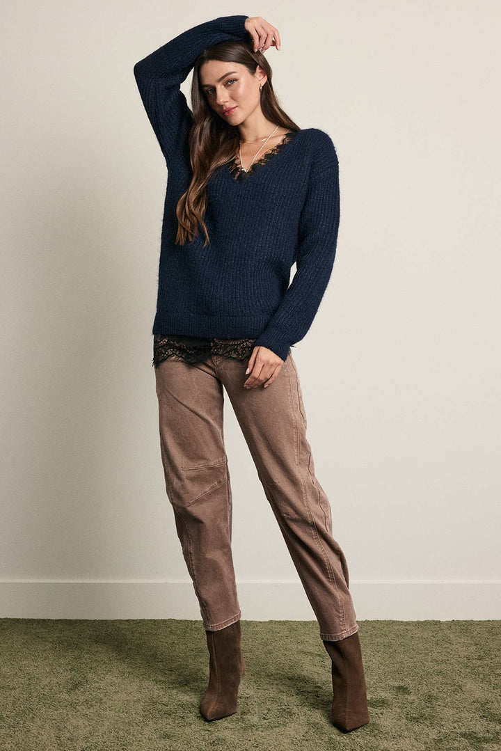 A woman modeling a navy blue ribbed knit sweater with a lace-trimmed neckline, paired with brown pants and brown ankle boots, standing against a plain beige wall.