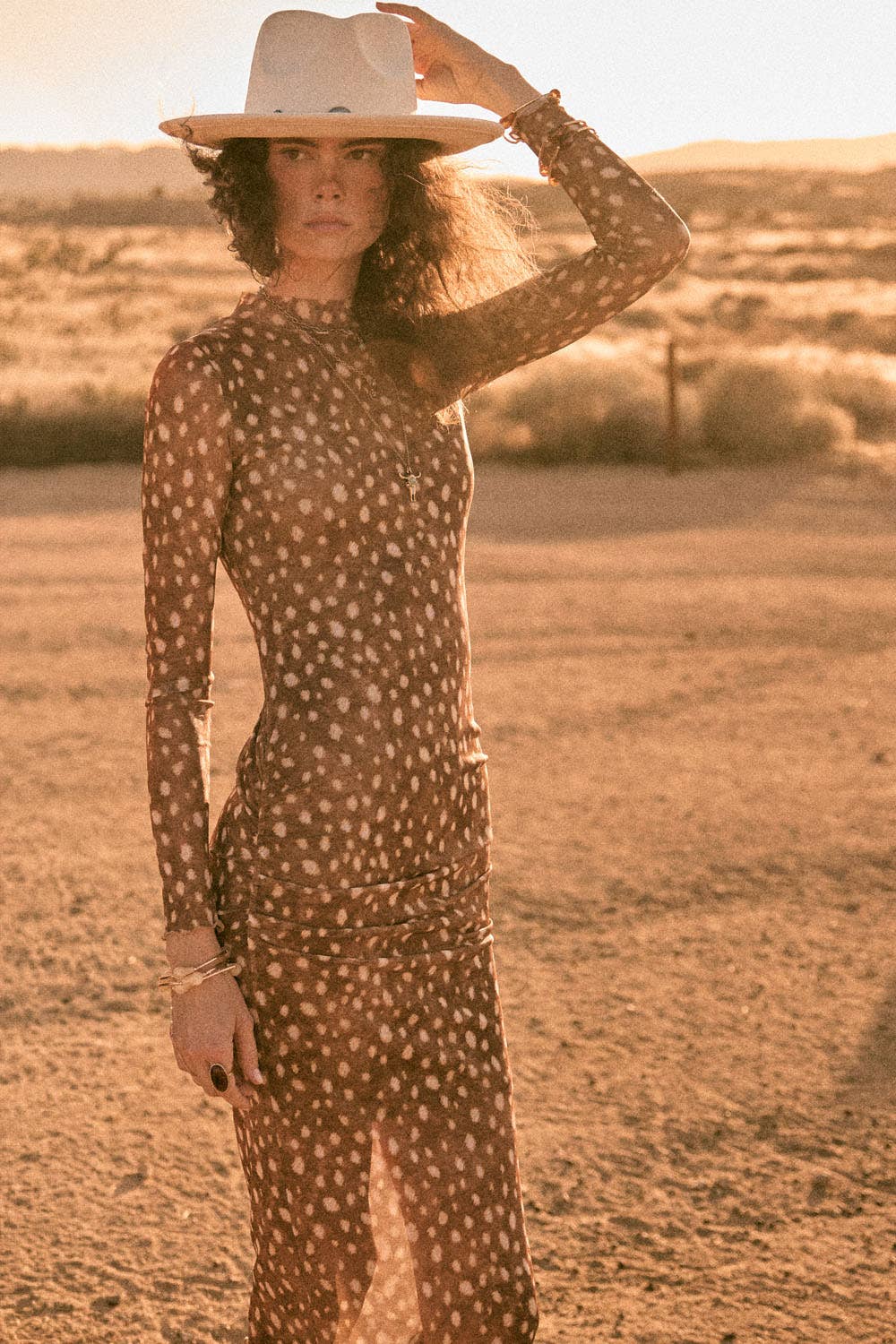 A woman stands in a desert landscape wearing a fitted long-sleeve midi dress with a small white dot pattern on a brown base, paired with a wide-brim white hat.