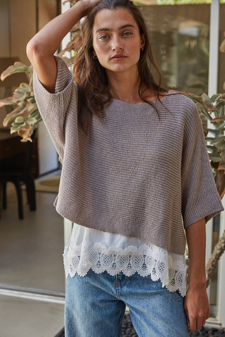 A woman wearing a brown ribbed knit short-sleeve sweater top with a white lace-trimmed hem, paired with blue jeans, standing in a softly lit indoor setting with plants in the background.