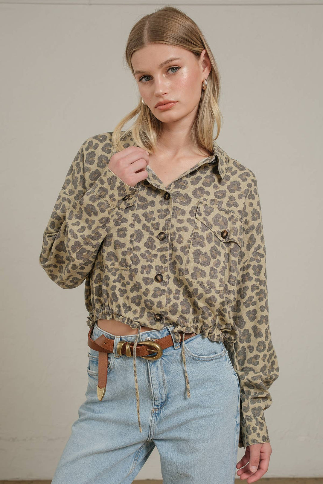 A woman wearing a beige and brown leopard-print button-up shirt with light-wash blue jeans with a brown belt, standing against a plain light beige wall.