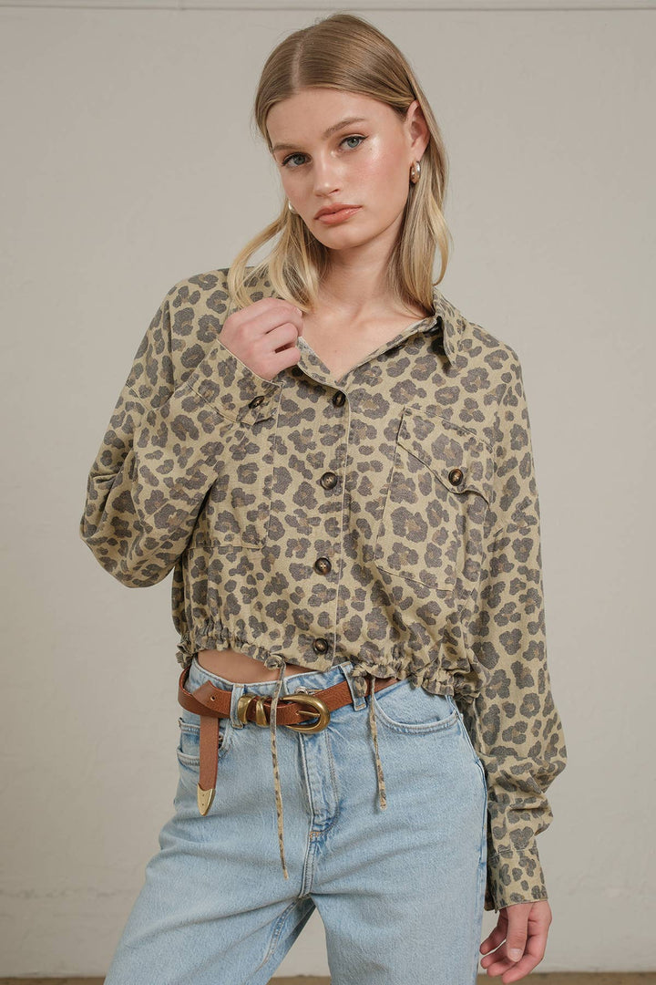 A woman wearing a beige and brown leopard-print button-up shirt with light-wash blue jeans with a brown belt, standing against a plain light beige wall.