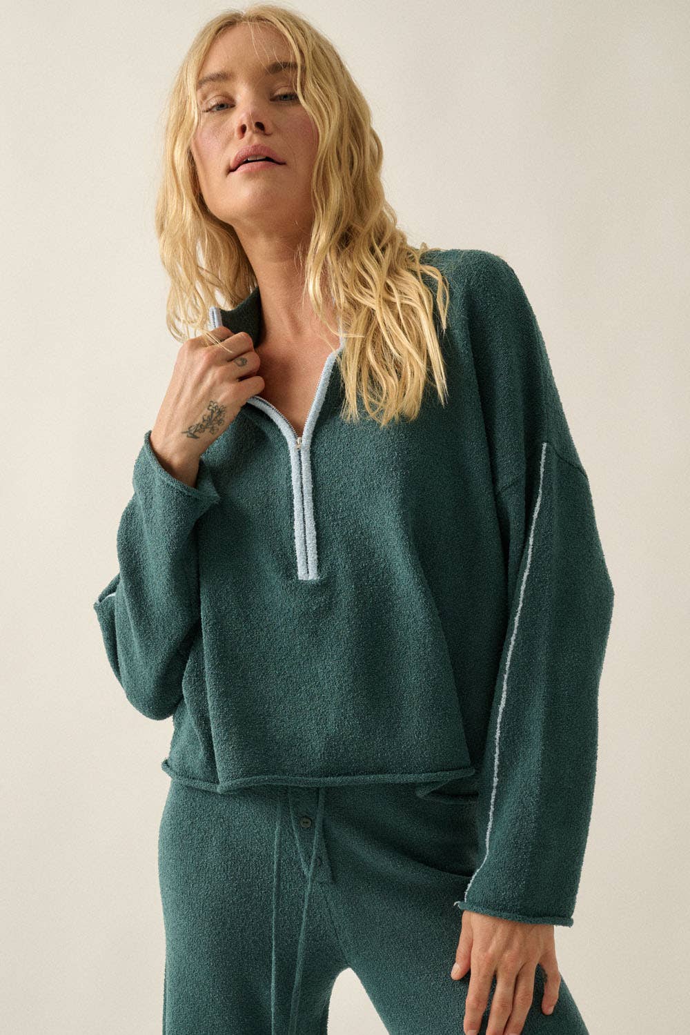 A woman wearing a matching dark teal sweater lounge set, featuring a cropped quarter-zip sweater and coordinating sweater pants, against a plain light beige background.
