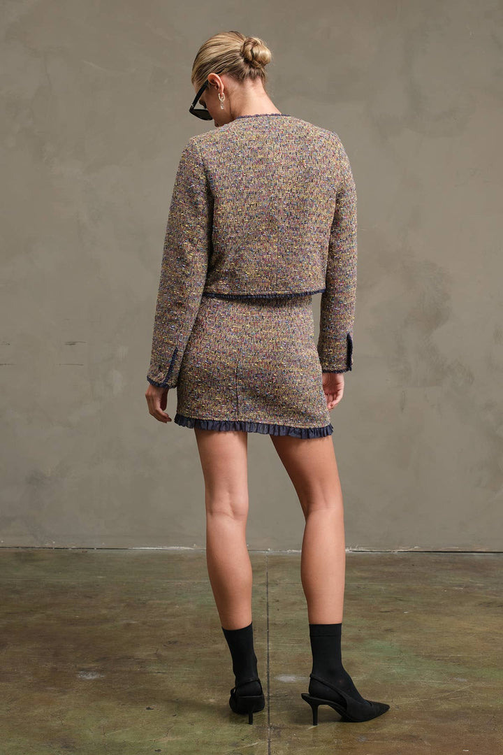 A woman photographed from behind wearing a tweed mini skirt with a cropped jacket-style top, black ankle socks, and black pointed-toe heels against a textured gray studio wall.