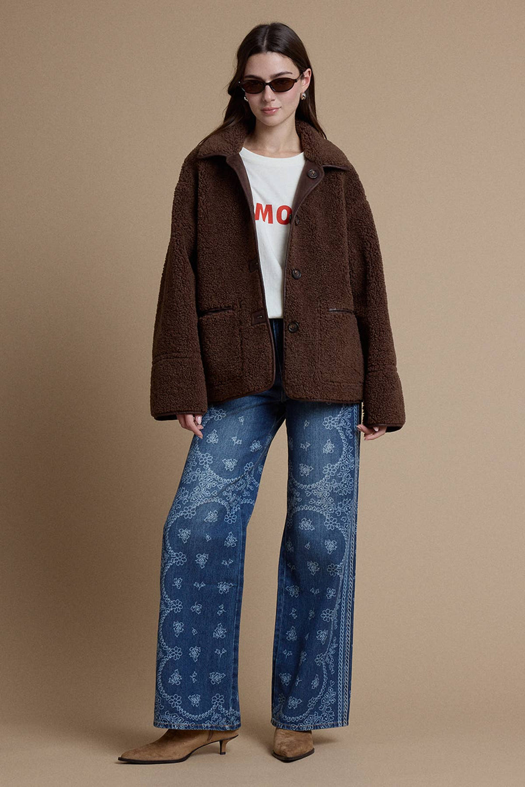 A model wearing a brown fuzzy teddy coat, a white graphic t-shirt, blue paisley wide-leg jeans, and brown heeled mules, standing against a solid beige studio background.