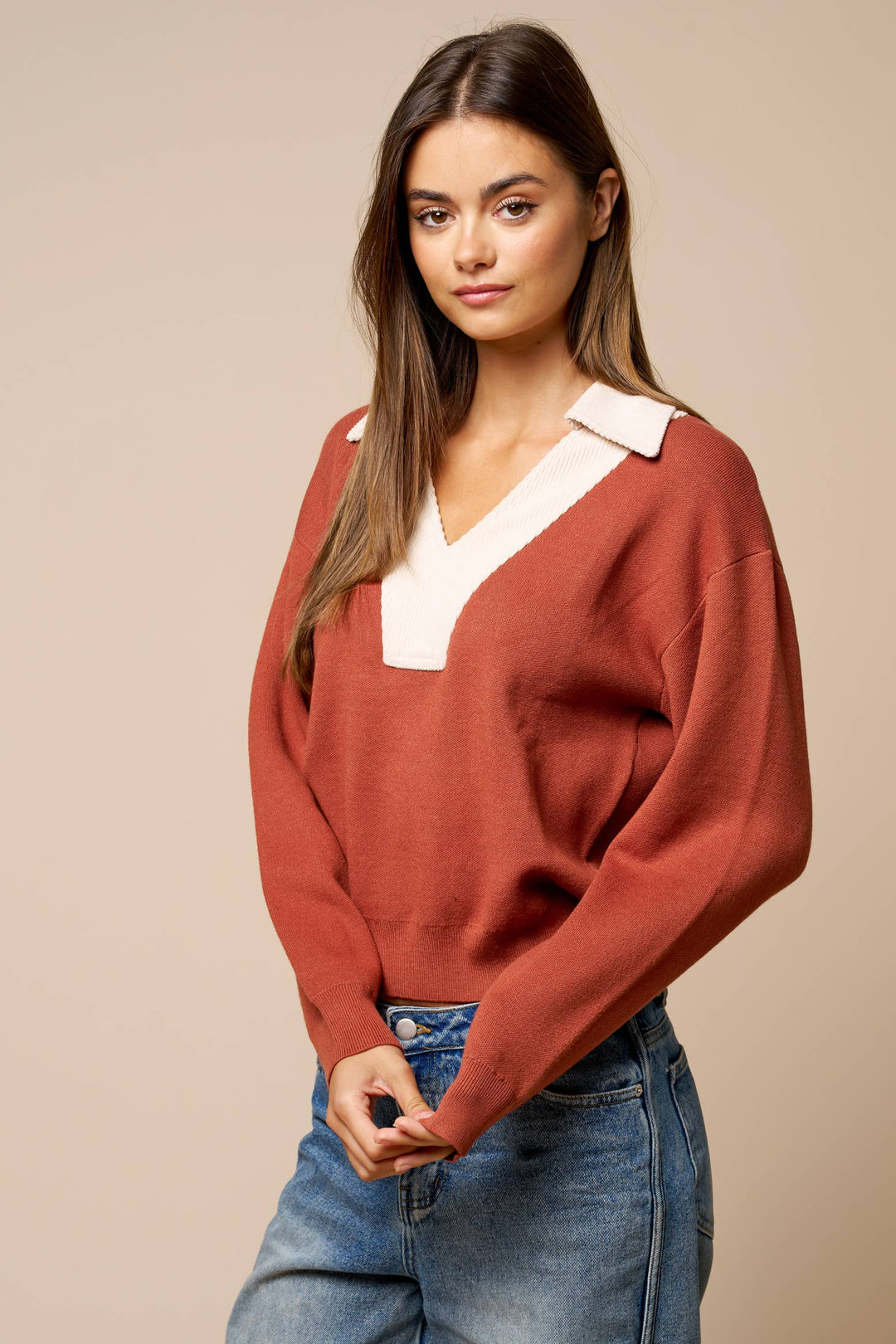 A woman wearing a rust-colored knit sweater with a contrasting white V-neck collar, paired with blue jeans, against a solid beige studio background.