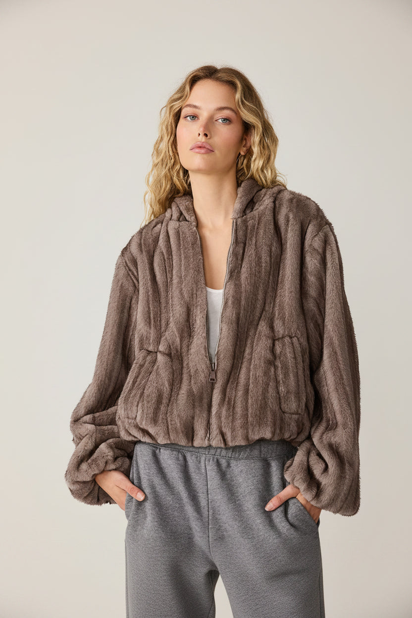 A woman wearing a brown faux fur zip-up hoodie jacket with a hood and front pockets, paired with gray sweatpants, shown against a plain white background.