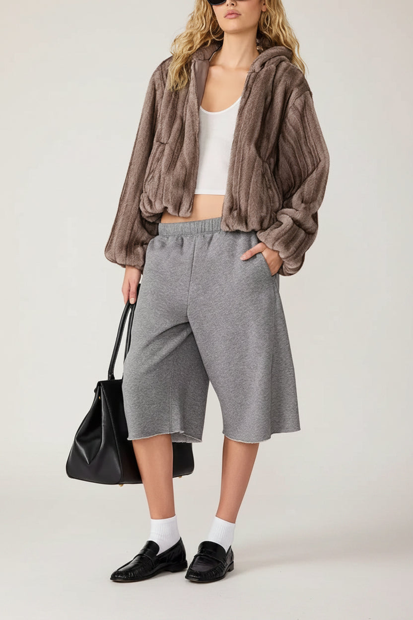A model wearing a brown fuzzy cropped hoodie, gray cropped sweatpants, a white tank top, black loafers, white socks, and carrying a black tote bag, set against a plain white background.