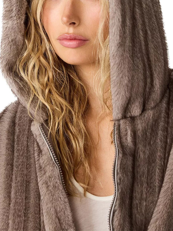 Close-up of a woman wearing a plush faux-fur zip hoodie in taupe brown, with her hood up and long blonde hair visible against a white background.