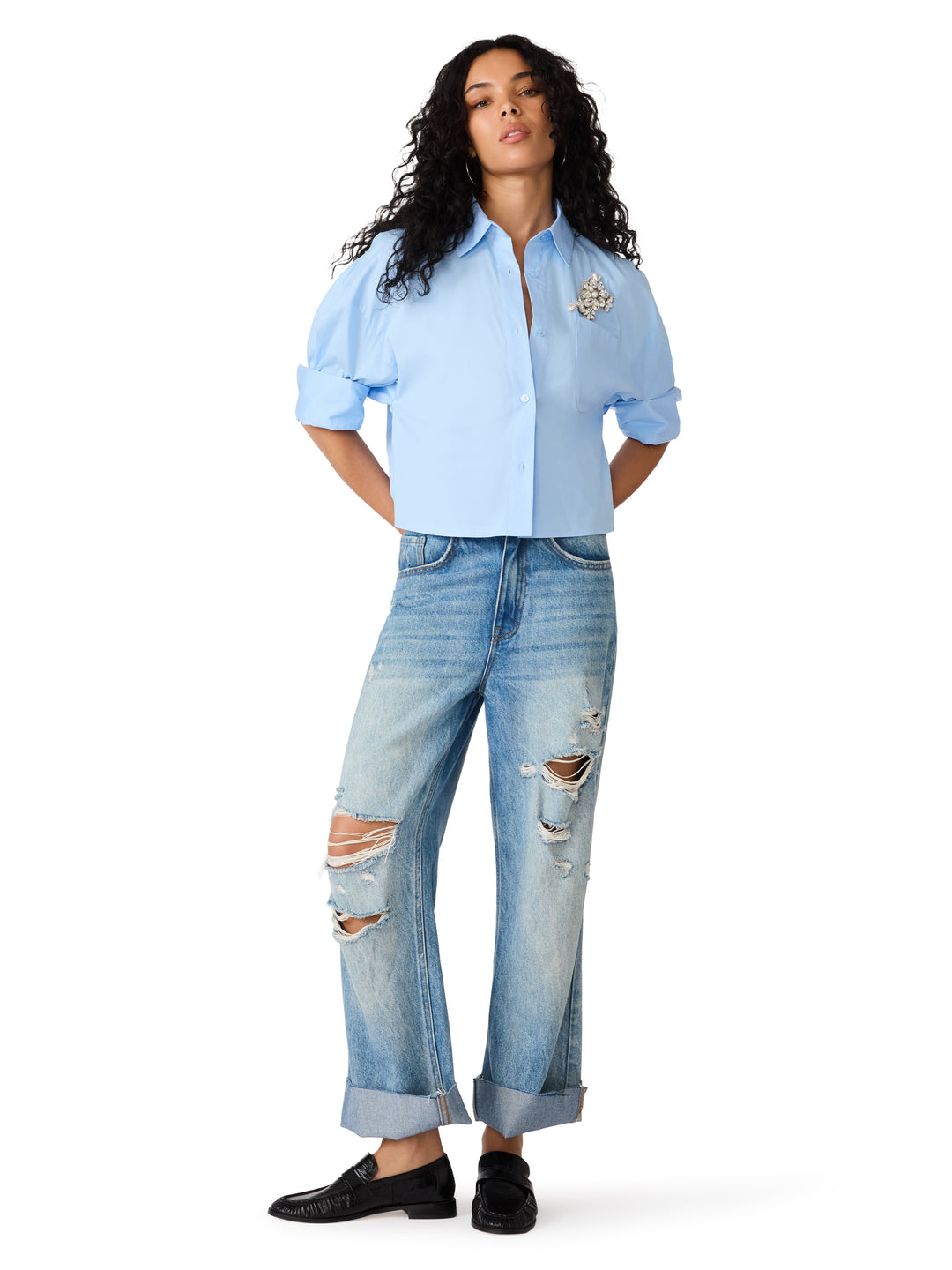 A model wearing a light blue cropped button-up shirt with rolled sleeves, distressed light-wash blue jeans, and black loafers, standing against a solid white background.