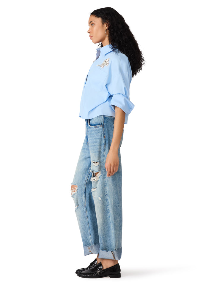 A model shown in profile wearing a light blue cropped button-up shirt with a small embroidered detail on the chest, light-wash distressed wide-leg jeans, and black leather loafers, set against a plain white background.
