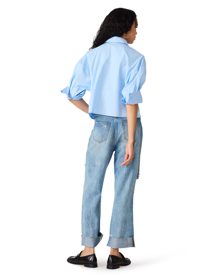 A model shown from the back wearing a light blue cropped button-up shirt with voluminous rolled sleeves, light-wash blue jeans, and black loafers, set against a plain white background.