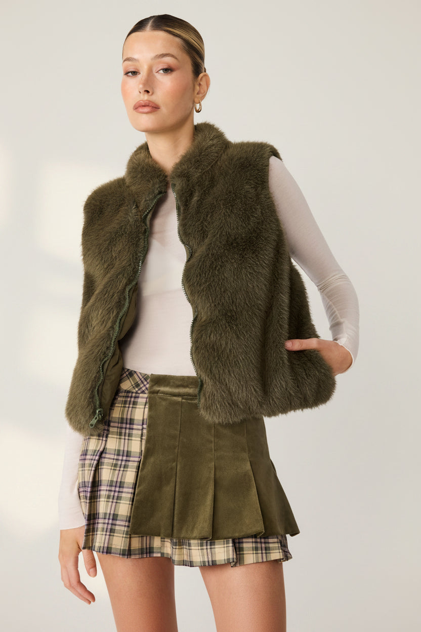 A model wearing an olive green faux fur vest over a cream long-sleeve top and a matching olive green pleated skirt with a plaid pattern, set against a plain white background.