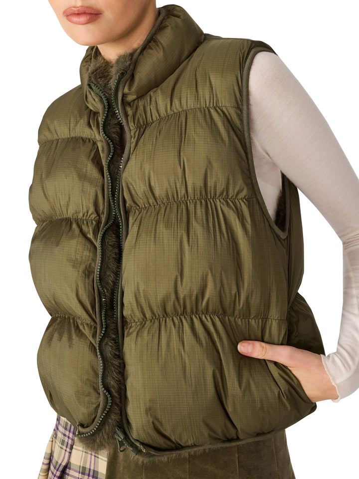 An olive green quilted puffer vest with a high collar and dark zipper, worn by a model over a white long-sleeve top.