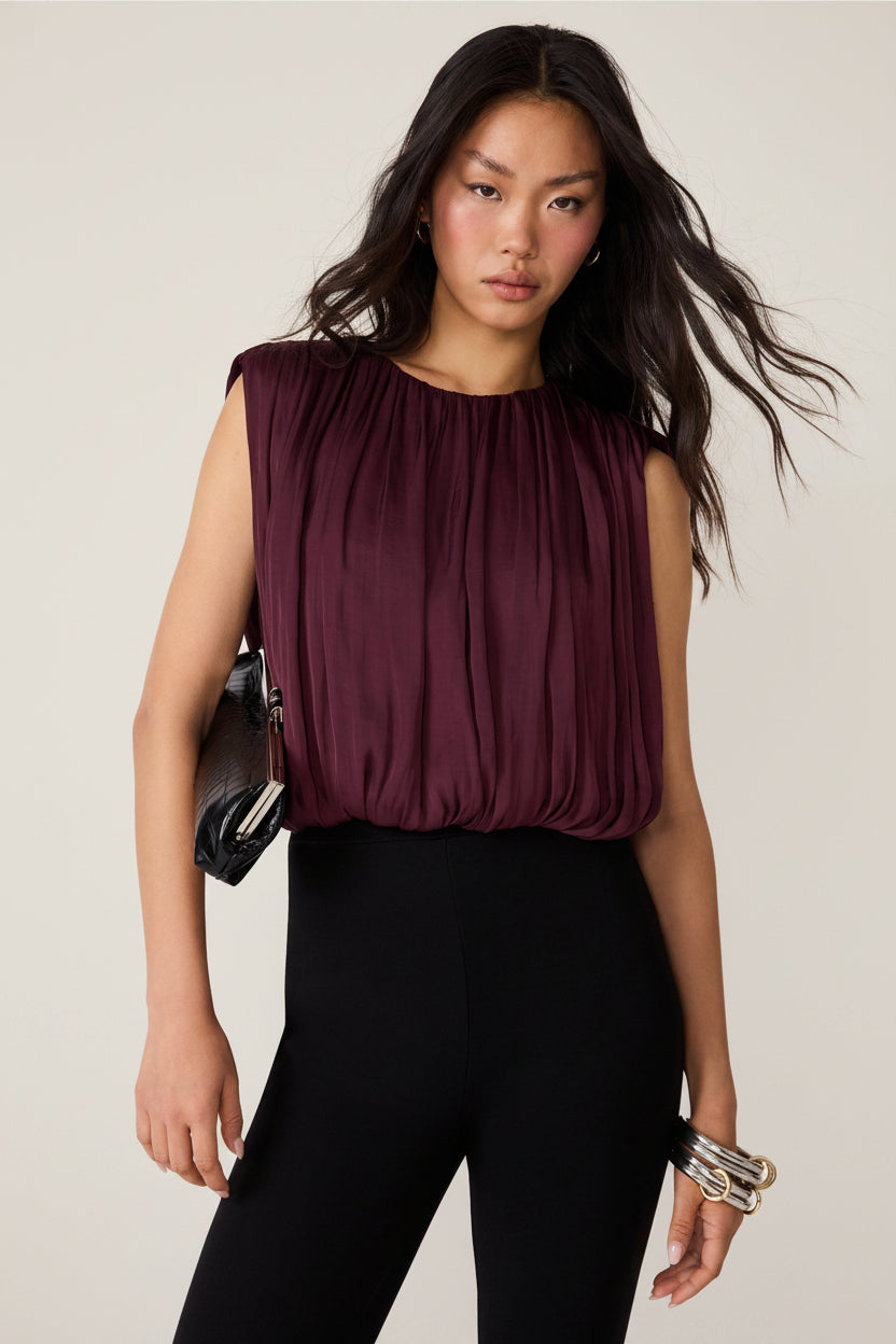 A woman modeling a sleeveless pleated top in deep burgundy, paired with black pants, a black clutch, and silver bracelets, against a plain white background.