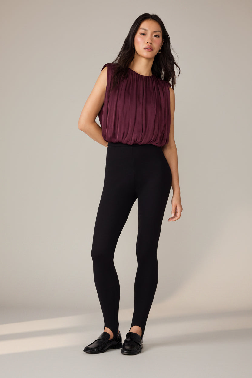 A woman modeling a sleeveless pleated top with bubble hem in deep burgundy and black leggings, paired with black loafers, against a plain white background.