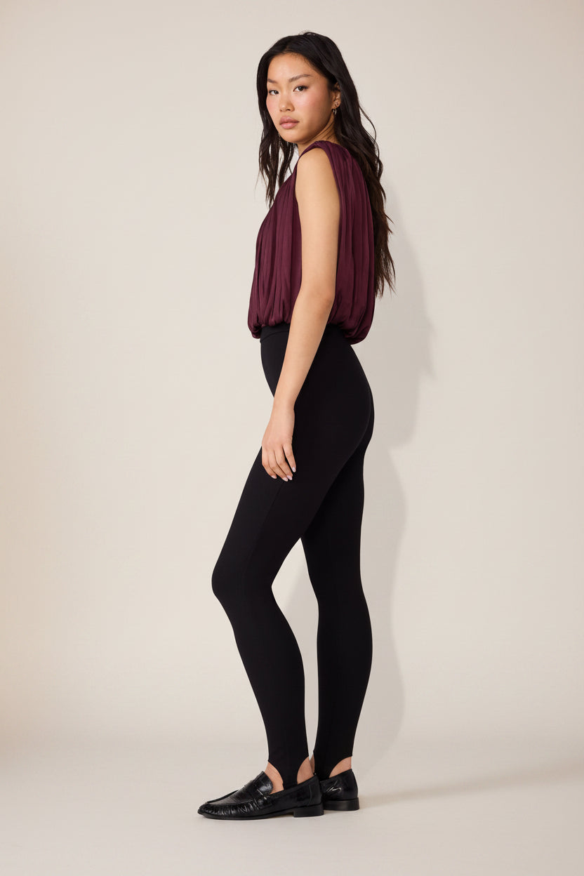 A woman modeling a sleeveless deep burgundy top and black leggings, standing in profile against a plain white studio background.