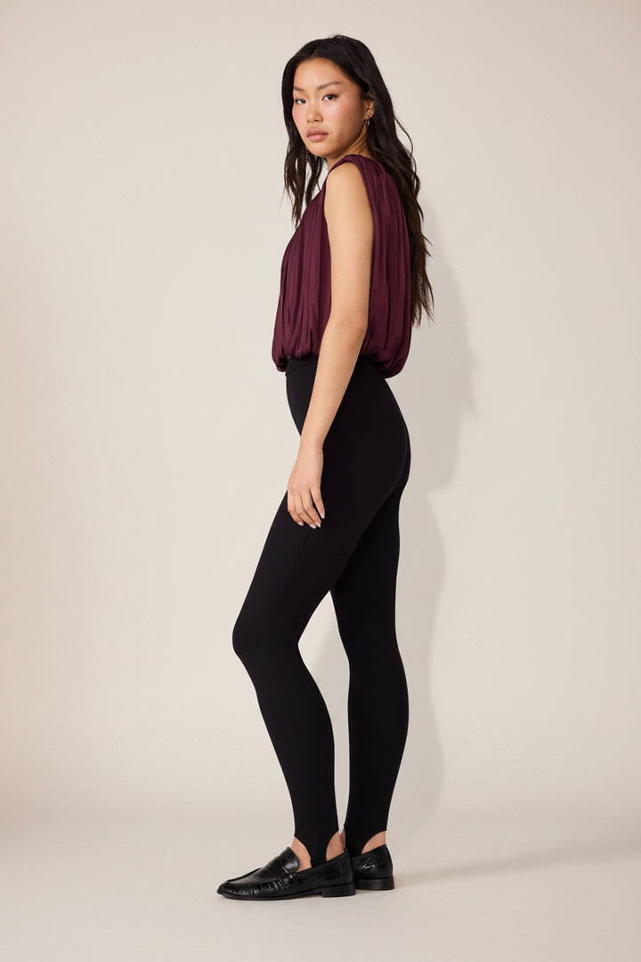 A woman modeling a sleeveless deep burgundy top and black leggings, standing in profile against a plain white studio background.