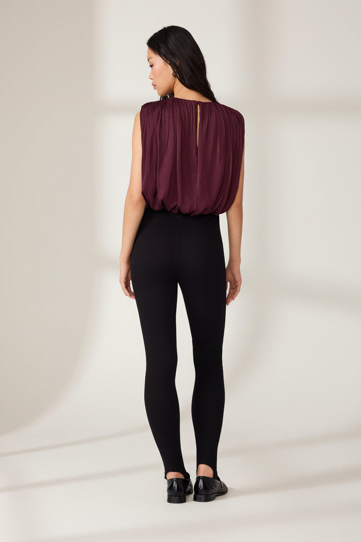 A woman shown from the back wearing a sleeveless deep burgundy top with a gathered, pleated back detail and black leggings, photographed against a plain white studio background.