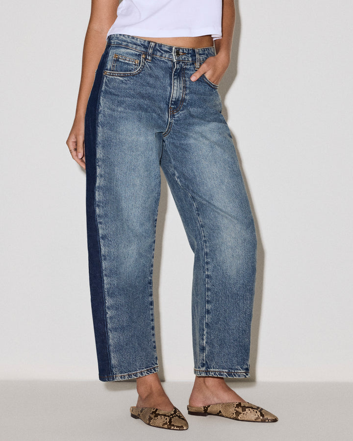 A model wearing cropped, barrel-leg blue denim jeans with a dark navy vertical stripe running down the left leg, paired with a white crop top and snakeskin-patterned flat shoes, against a plain light gray studio backgroung