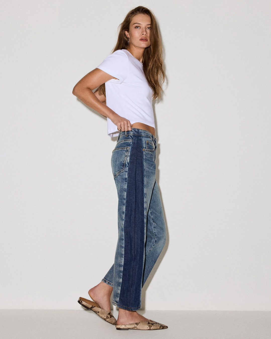 A woman modeling a pair of barrel-leg blue jeans with a dark navy vertical stripe down the front, styled with a white cropped t-shirt and flat sandals against a plain light gray studio background.