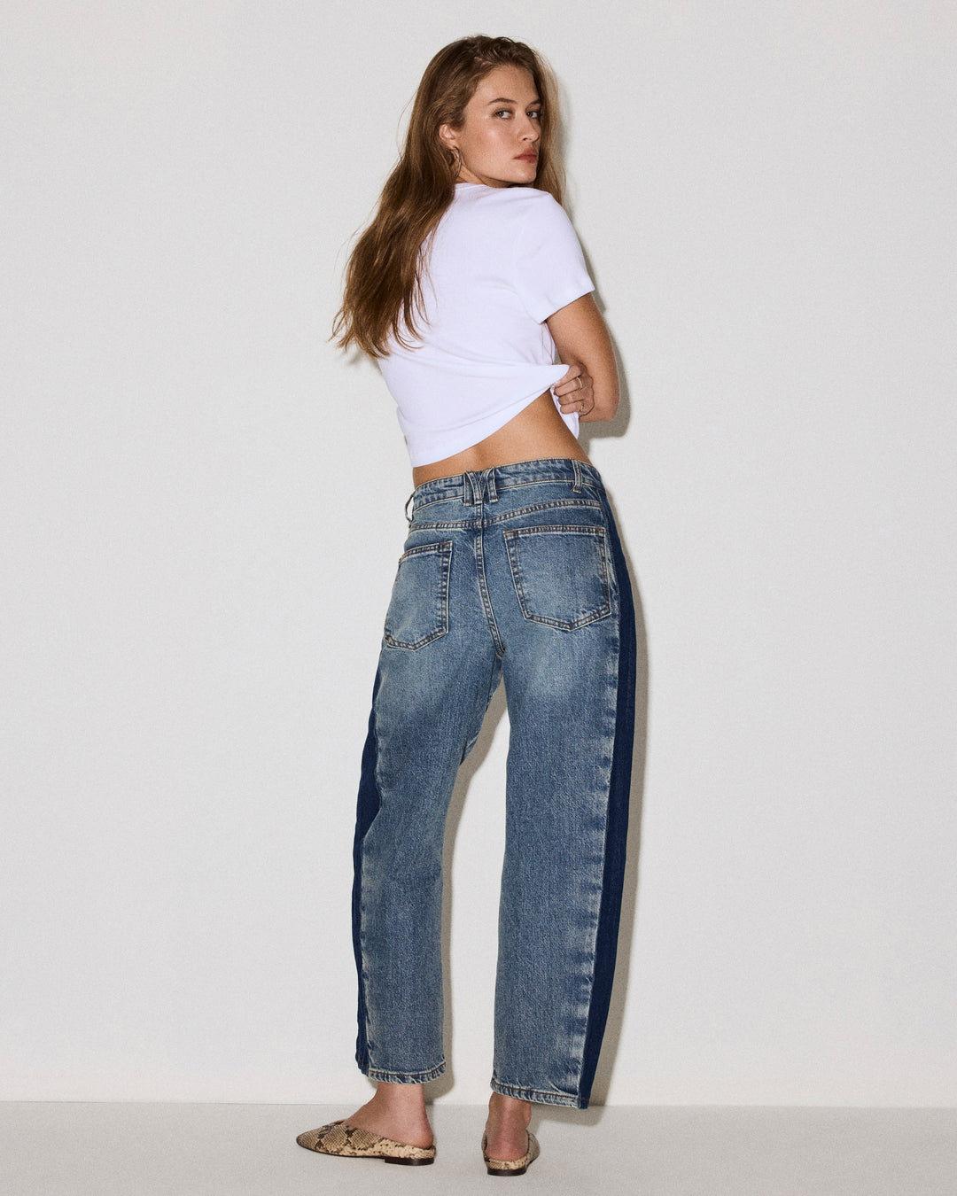 A woman wearing high-waisted, wide-leg blue jeans with dark navy side stripes, styled with a white cropped t-shirt and leopard print flat shoes, photographed against a plain white studio background.