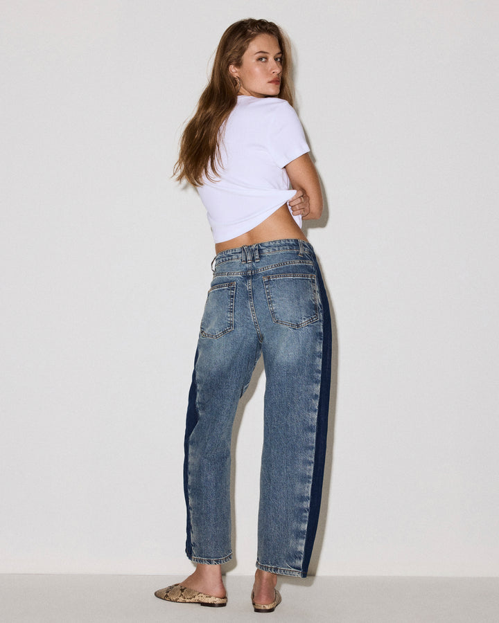 A woman wearing high-waisted, wide-leg blue jeans with dark navy side stripes, styled with a white cropped t-shirt and leopard print flat shoes, photographed against a plain white studio background.