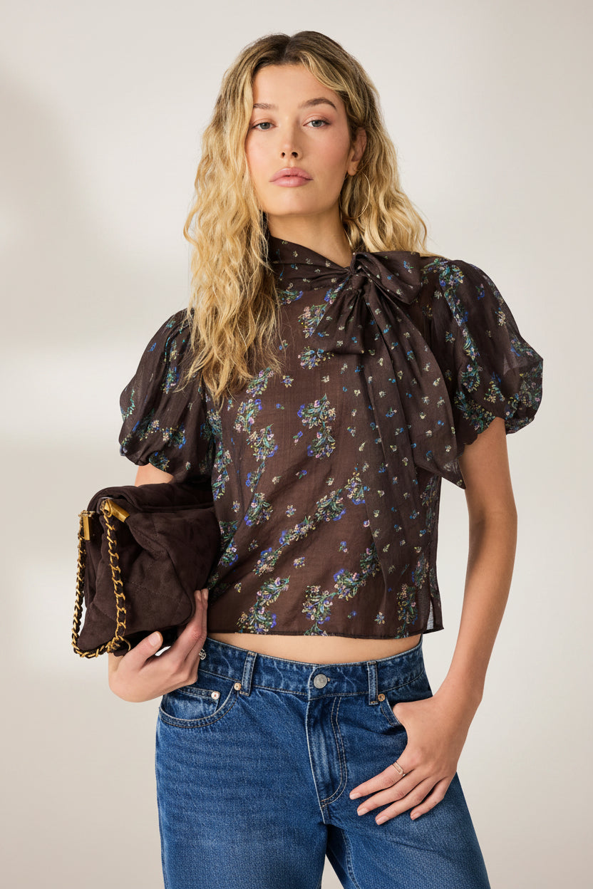 A woman modeling a dark brown sheer puff-sleeve blouse with a blue and yellow floral print, a large bow at the collar, and blue jeans, holding a matching brown clutch bag with a gold chain strap against a plain white background.