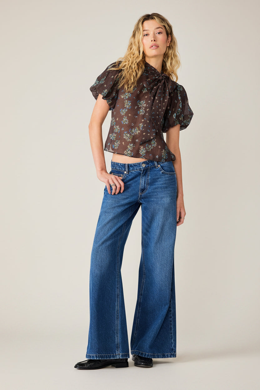A model stands against a white studio background wearing a dark brown short-sleeve blouse with a blue and white floral pattern, paired with high-waisted blue flare jeans and black shoes.