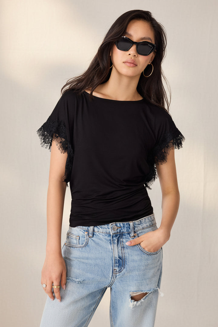 A woman modeling a black short-sleeve top with ruffled lace trim at the sleeves, paired with light blue distressed jeans and black sunglasses, against a plain white background.