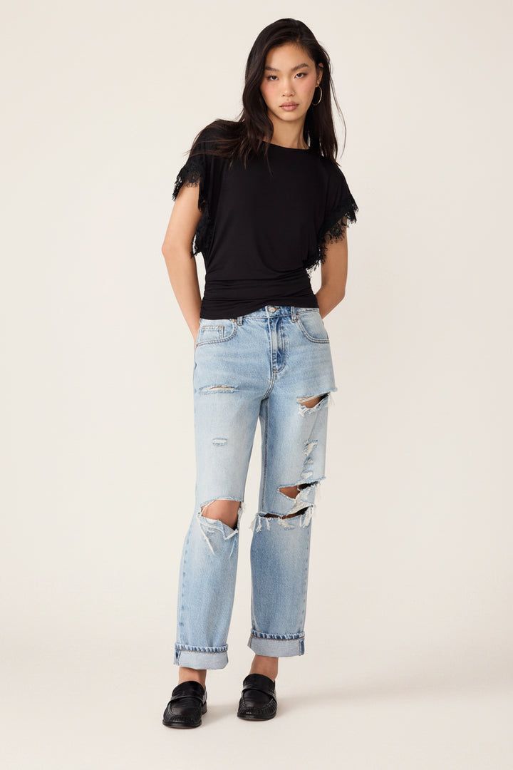 A model wearing a black short-sleeve top with ruffled shoulders and light-wash distressed jeans, standing against a plain white background.