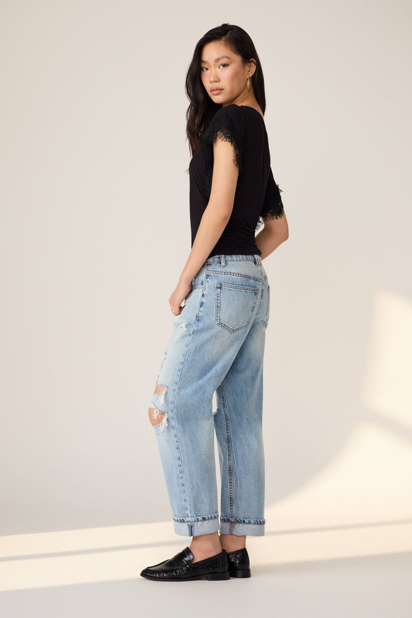 A model shown from the back wearing light-wash distressed jeans with ripped knees, a black sleeveless top with lace hem detail, and black loafers, against a plain white background.