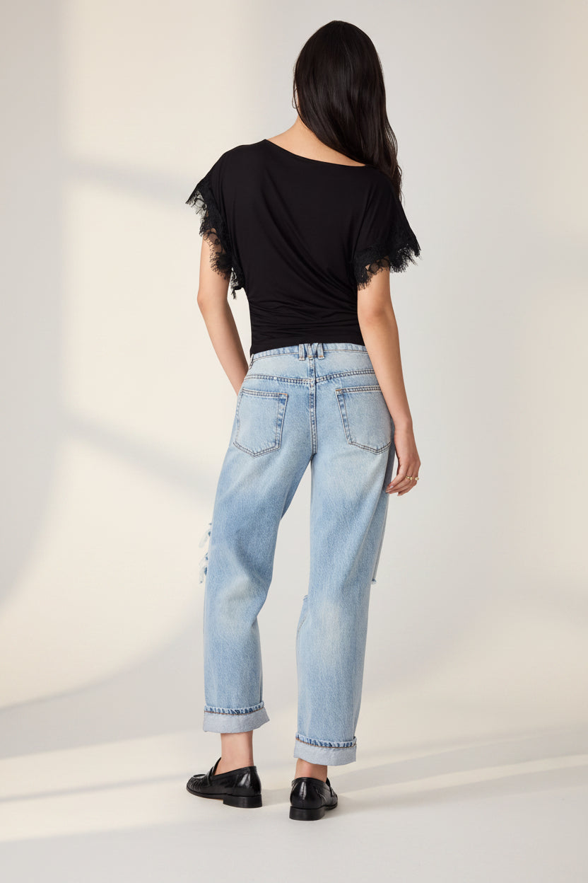 A woman shown from the back wearing a black short-sleeve top with lace trim, light-wash blue jeans with rolled cuffs and distressed details, and black shoes, against a plain white background.