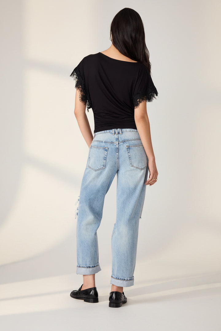 A woman shown from the back wearing a black short-sleeve top with lace trim, light-wash blue jeans with rolled cuffs and distressed details, and black shoes, against a plain white background.