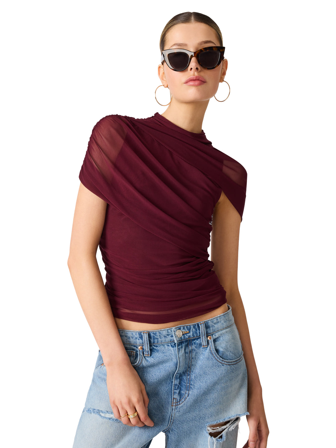 A woman modeling a ruched, short-sleeve top in deep burgundy with sheer mesh side panels, paired with light-wash distressed jeans and black sunglasses against a plain white background.