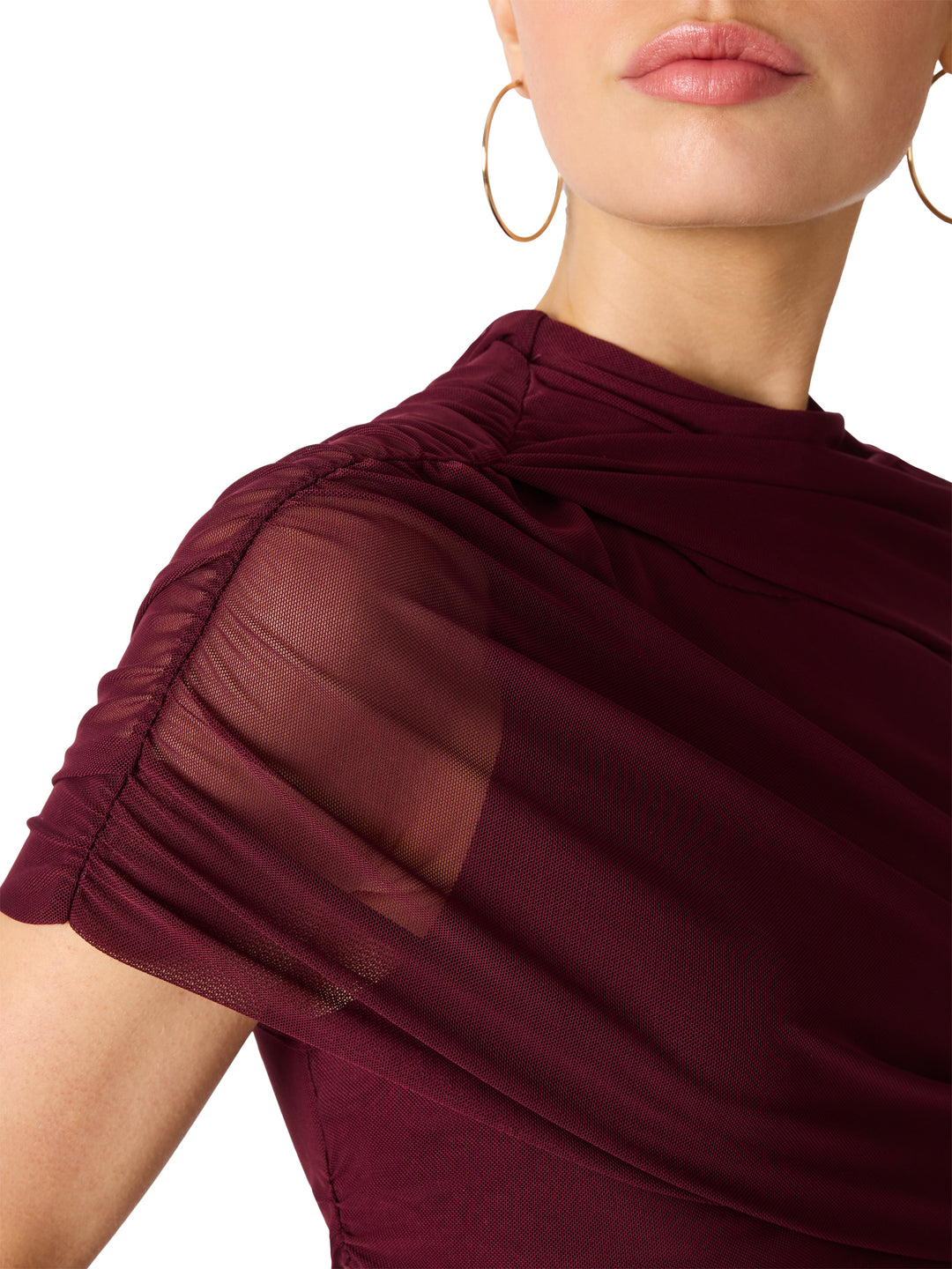 Close-up of a model wearing a draped, short-sleeved top in deep burgundy with sheer mesh detailing on the shoulder, paired with gold hoop earrings against a white background.