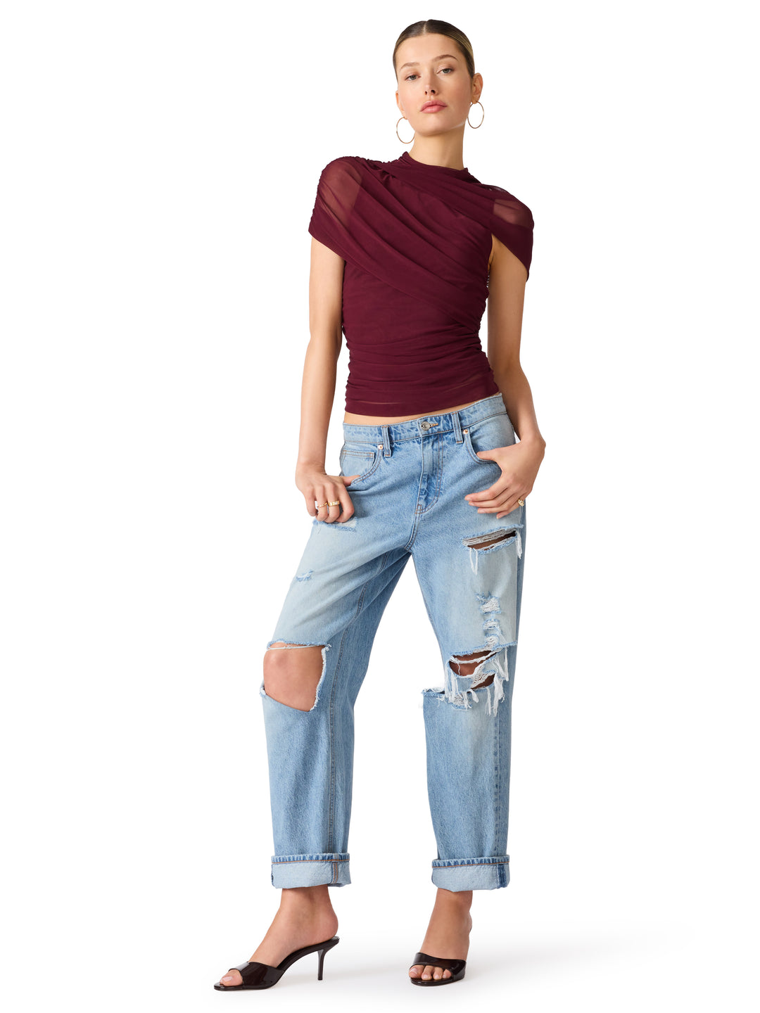 A model wearing a dark burgundy draped short-sleeve top, light-wash distressed jeans with ripped knees, and black heeled sandals, standing against a plain white background.