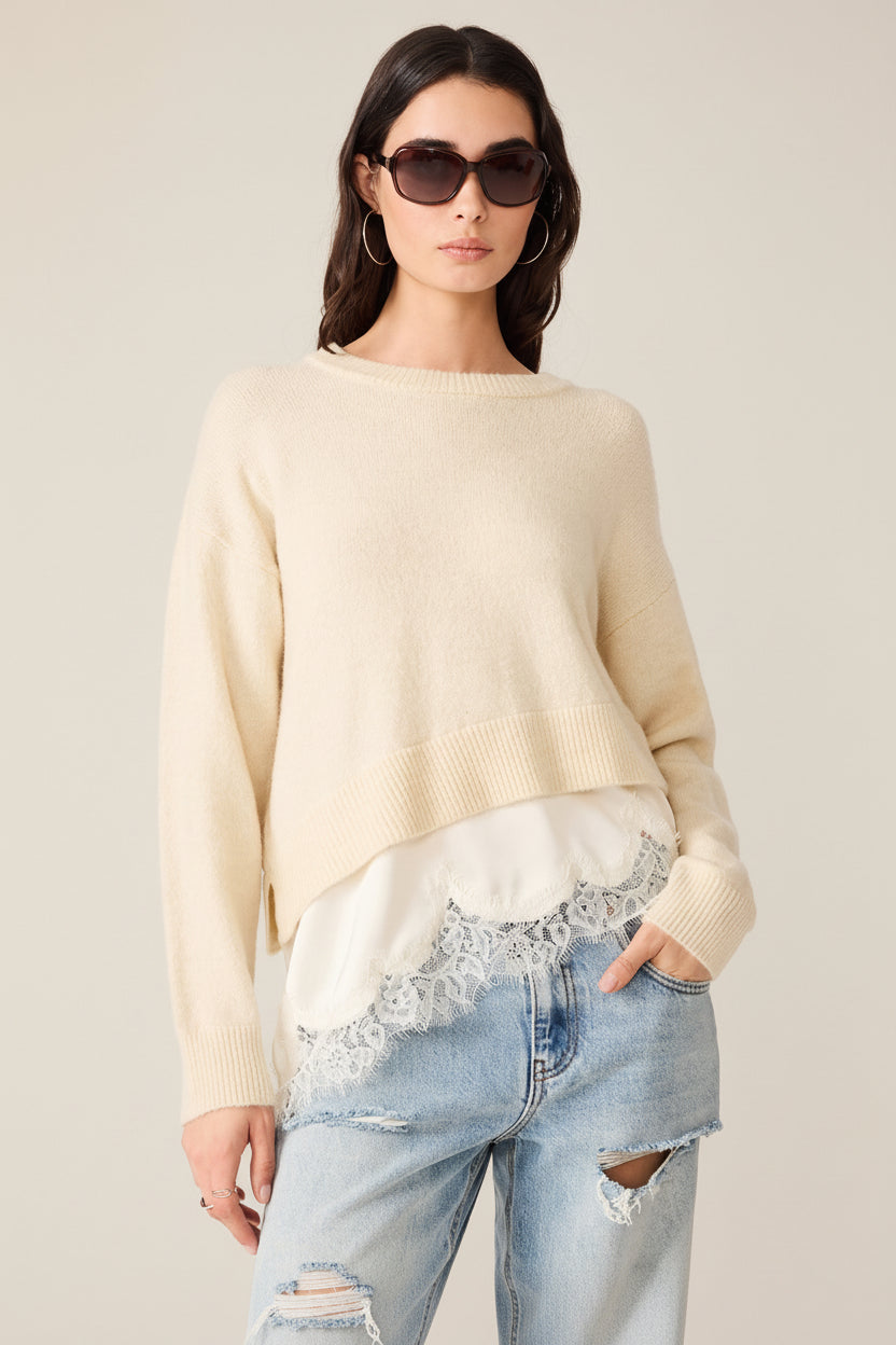 A woman modeling a cream-colored knit sweater with a layered white lace-trim hem, paired with light-wash distressed jeans and dark sunglasses, against a plain white background.