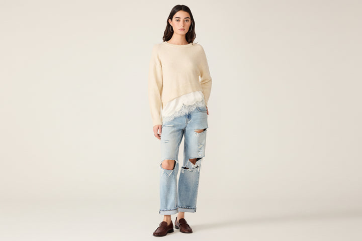 A model stands against a white studio background wearing a cream knit sweater with a white lace underlay  peeking out at the hem, light-wash distressed jeans with ripped knees, and dark brown loafers.