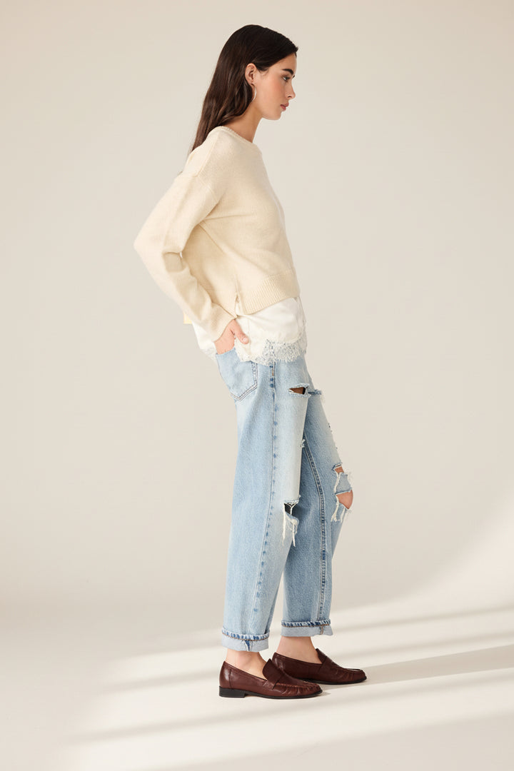 A woman shown in profile wearing a cream long-sleeve knit top, light-wash distressed jeans with ripped details, and dark brown leather loafers, photographed against a plain white studio background.