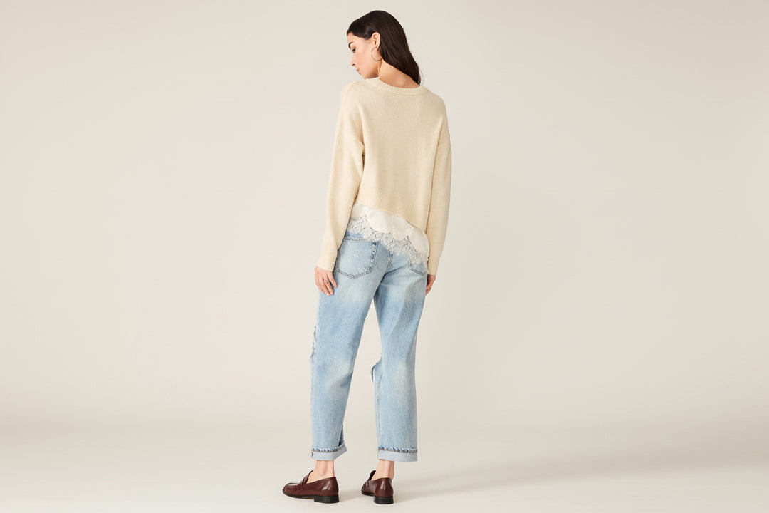 A woman shown from the back wearing a cream knit sweater with lace trim, light-wash blue jeans with lace detailing at the back hem, and dark brown loafers, against a plain white background.