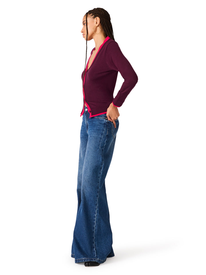 A woman shown in profile wearing a dark burgundy long-sleeve cardigan with a bright pink trim and blue flare jeans, standing against a plain white background
