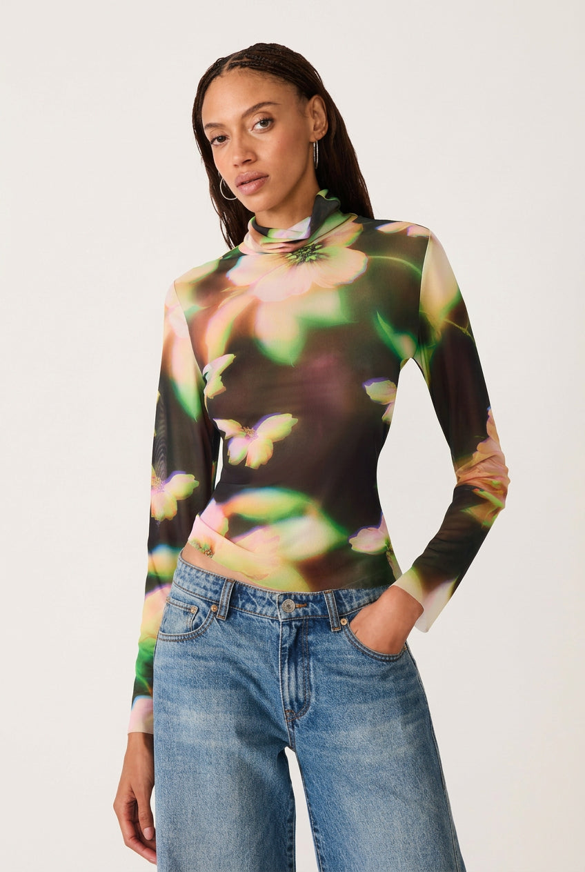 A woman modeling a sheer mesh long-sleeve mock neck top with a vibrant multicolor floral and butterfly print, paired with blue jeans, against a solid white background.
