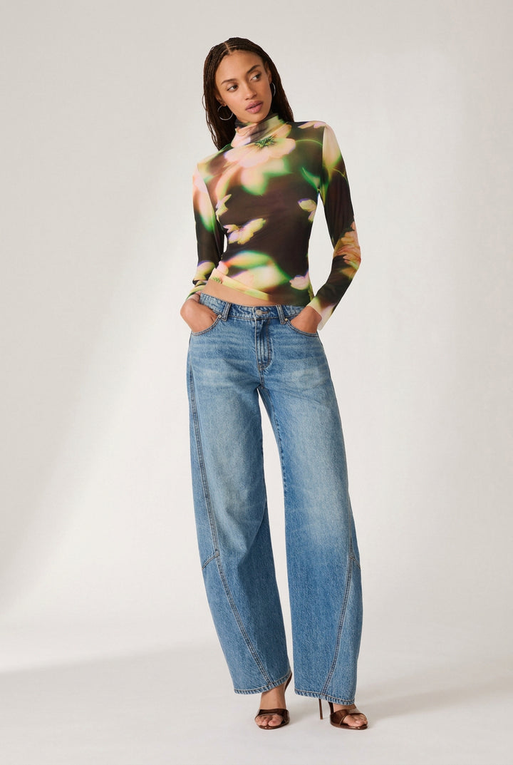 A model stands against a white studio background wearing a fitted long-sleeve mock-neck top with a multicolor abstract print in green, yellow, orange, and black, paired with wide-leg light-wash blue jeans and brown heeled sandals.
