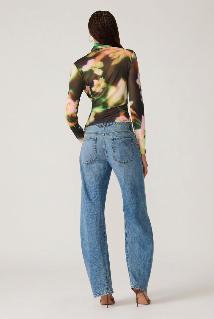 A woman shown from the back wearing a fitted long-sleeve top with a colorful abstract floral print in green, orange, pink, and black, paired with light blue jeans and brown heels, against a plain white studio background.
