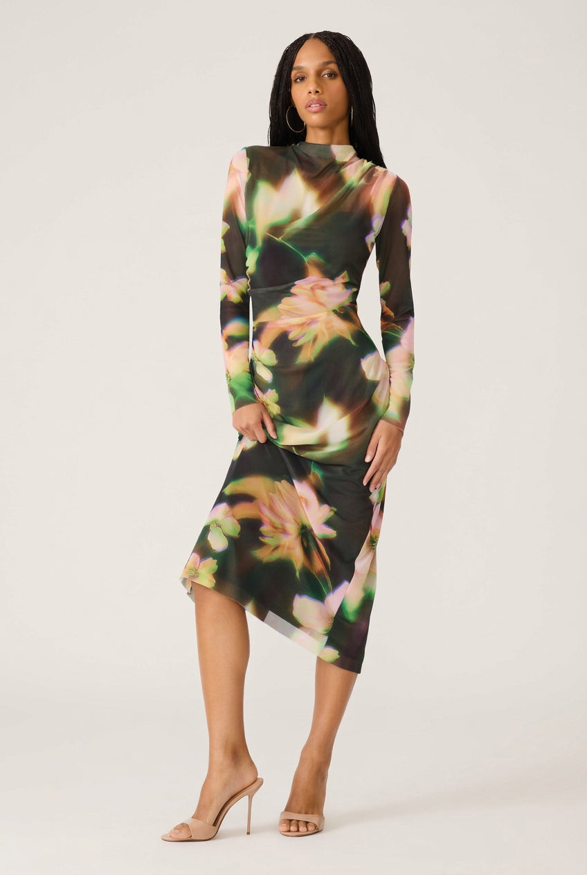 A woman modeling a long-sleeve midi dress with a dark base and a colorful floral print in pink, green, and yellow tones, paired with nude high-heeled sandals against a plain white background.