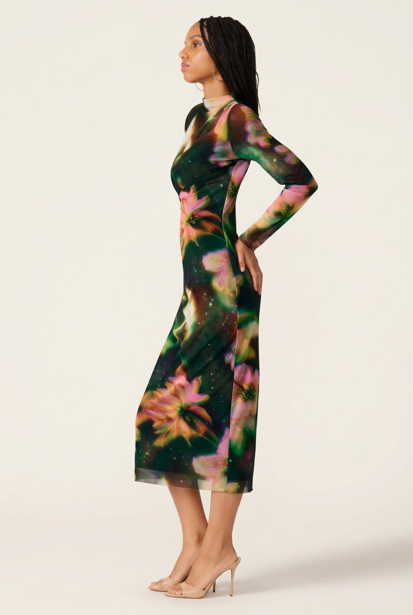 A woman modeling a long-sleeve mesh midi dress with a dark green base and a vibrant floral print in pink, yellow, and green, shown from a side profile against a white background.