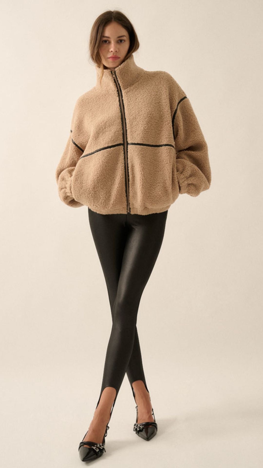 A woman wearing a beige sherpa fleece zip-up jacket with black faux leather contrast piping and black leather-look pants, photographed against a plain light beige studio background.