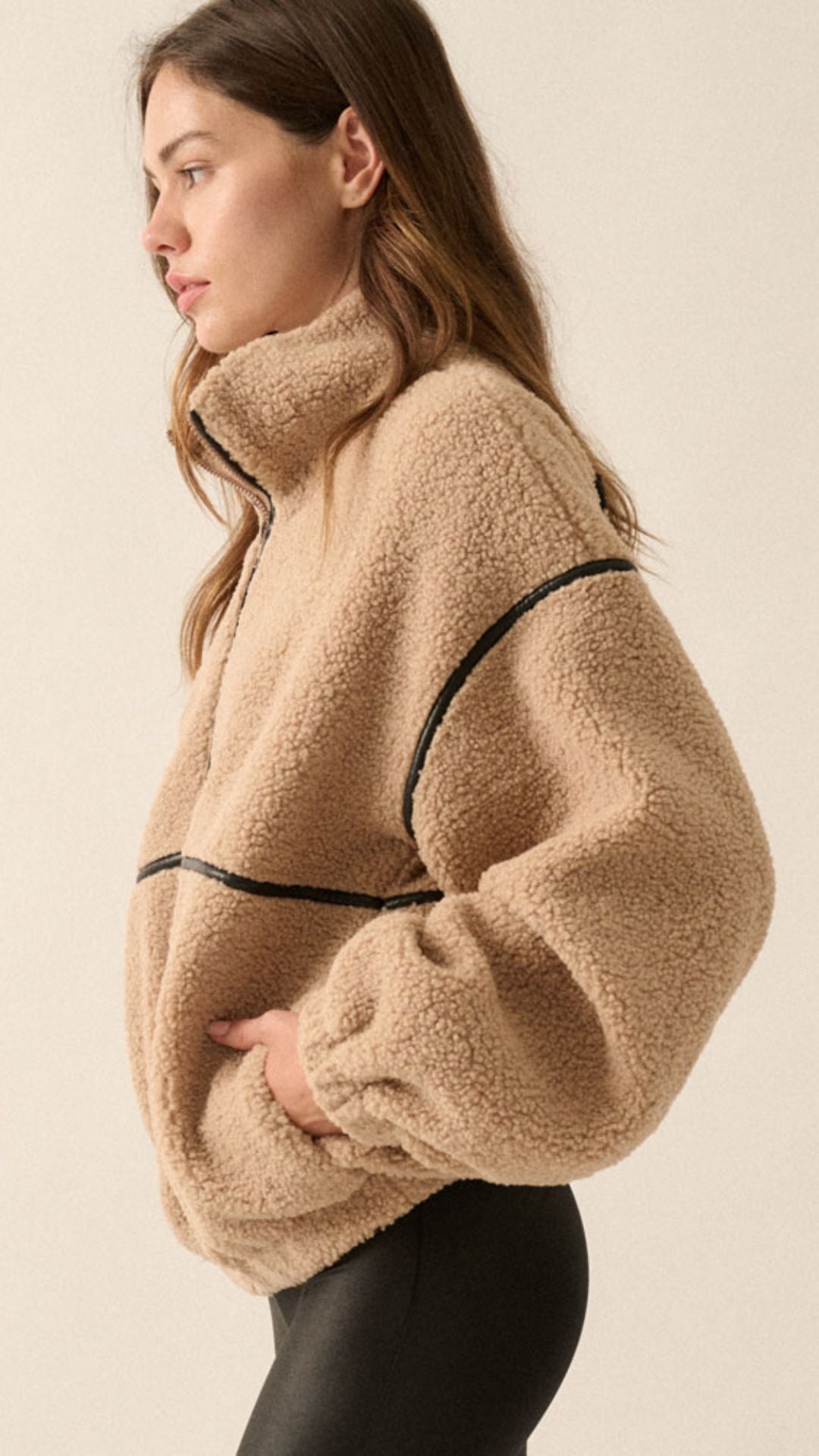 A woman wearing a beige sherpa fleece zip-up jacket with black faux leather contrast piping and black leather-look pants, photographed against a plain light beige studio background.