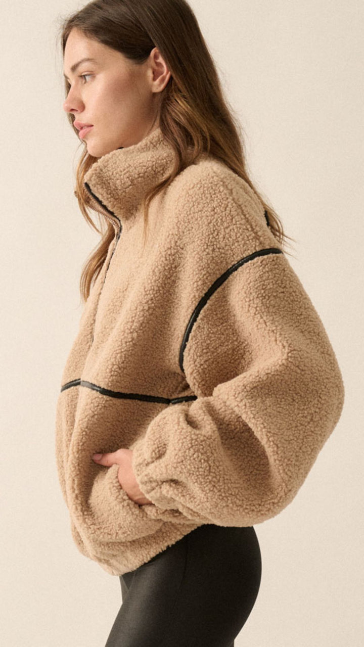A woman wearing a beige sherpa fleece zip-up jacket with black faux leather contrast piping and black leather-look pants, photographed against a plain light beige studio background.