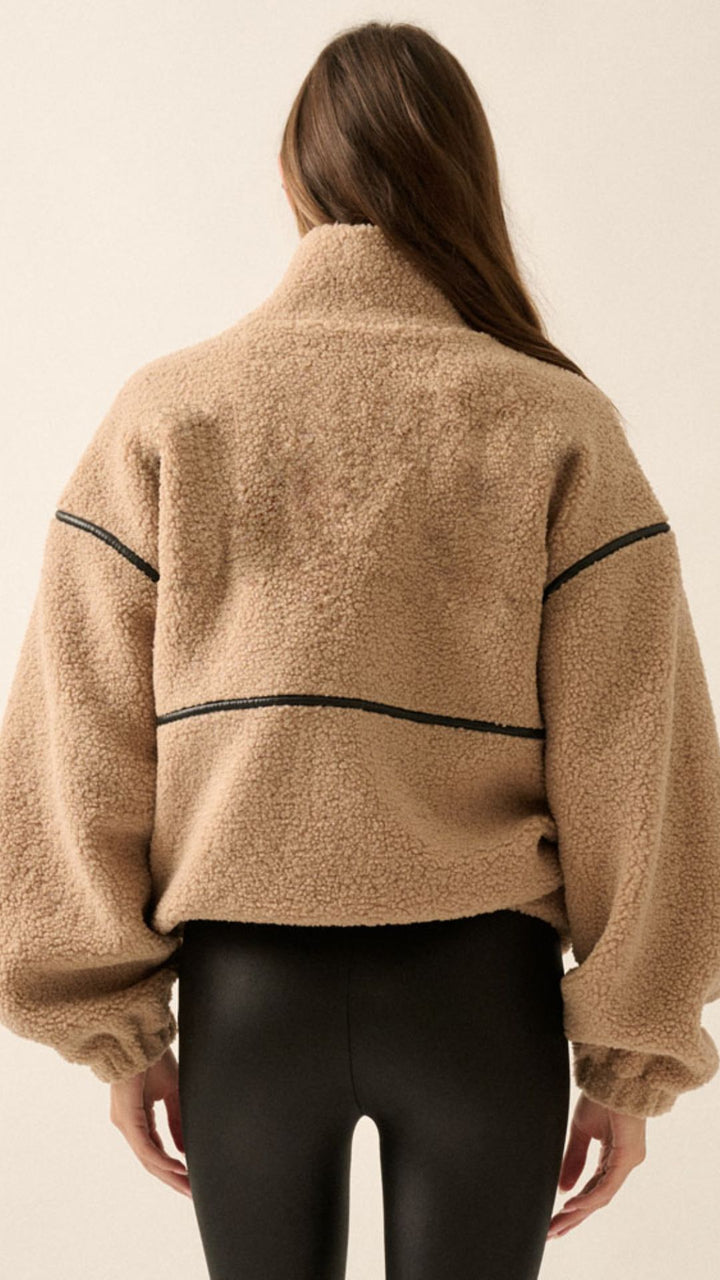 A woman wearing a beige sherpa fleece zip-up jacket with black faux leather contrast piping and black leather-look pants, photographed against a plain light beige studio background.